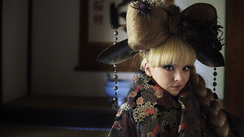 [photo] Close up of an actress in traditional Japanese headgear and attire standing infront of Japanese art indoors