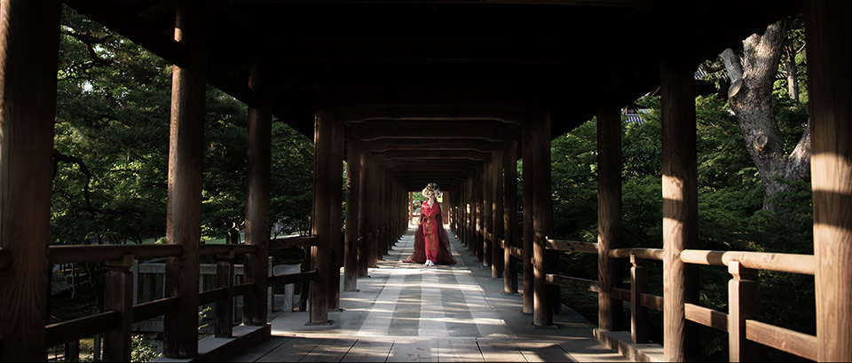 [photo] A color saturated version of a lady in traditional Japenese attire standing under a wooden bridge