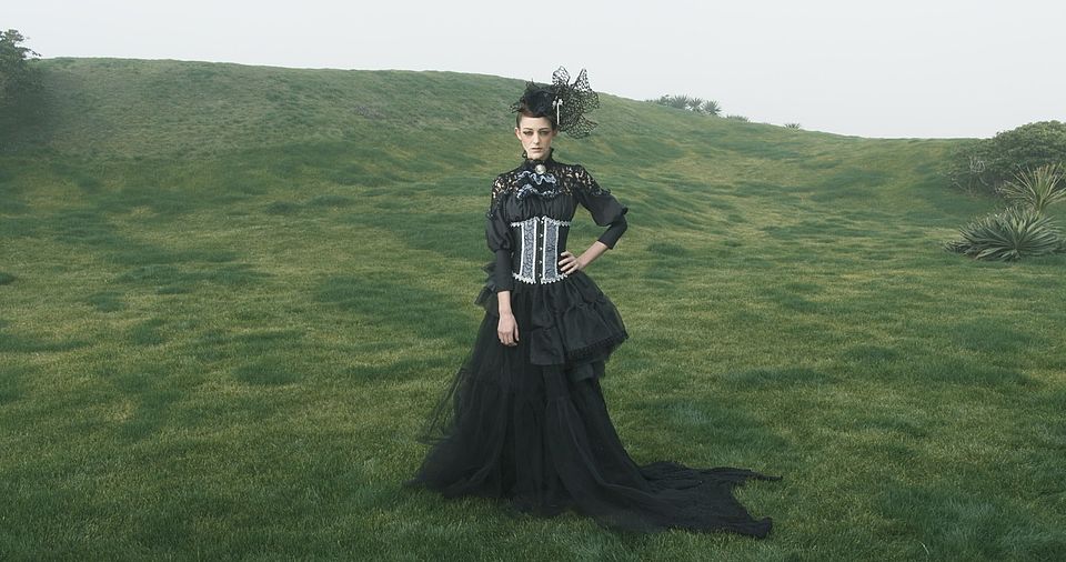 [photo] A wide shot of a lady dressed in a mediaval dress & hat standing on a green grass field on a cloudy day