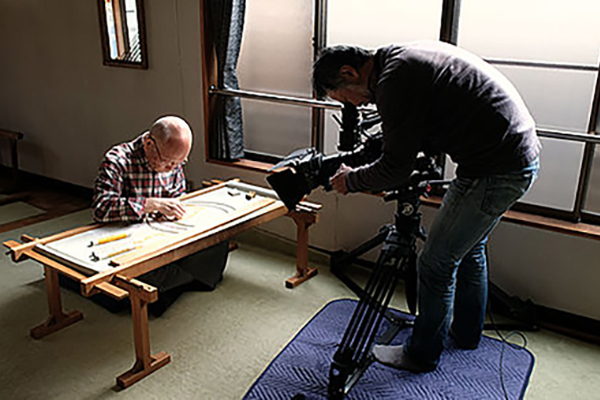 [photo] A cameraman shooting an older gentleman working with rulers on a table