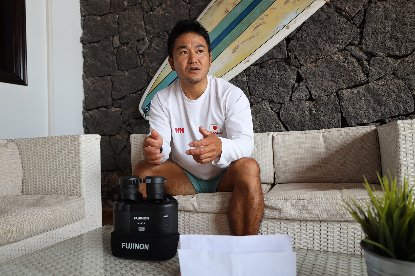 A short-haired man is sitting on a sofa, answering questions in an interview. On the table in front of him, there are binoculars.