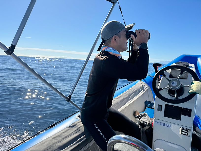 A man wearing a hat is looking into the distance through binoculars from a boat out at sea.