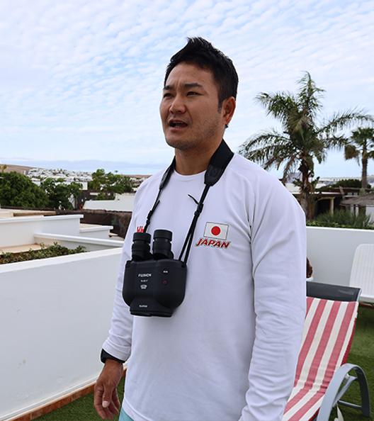 On the rooftop of a building, a short-haired man with binoculars around his neck is answering questions in an interview.