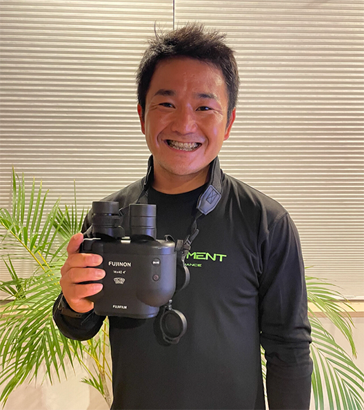 A short-haired man is holding binoculars and laughing.