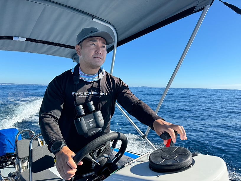 A man wearing a hat is steering a boat with binoculars around his neck.