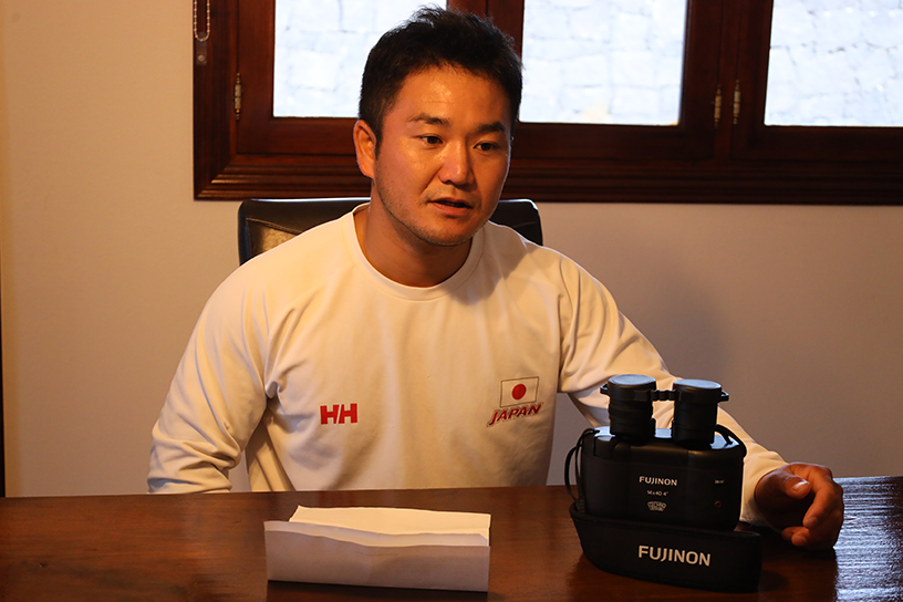 A short-haired man is sitting in a chair, answering questions in an interview. On the desk in front of him, there are binoculars.